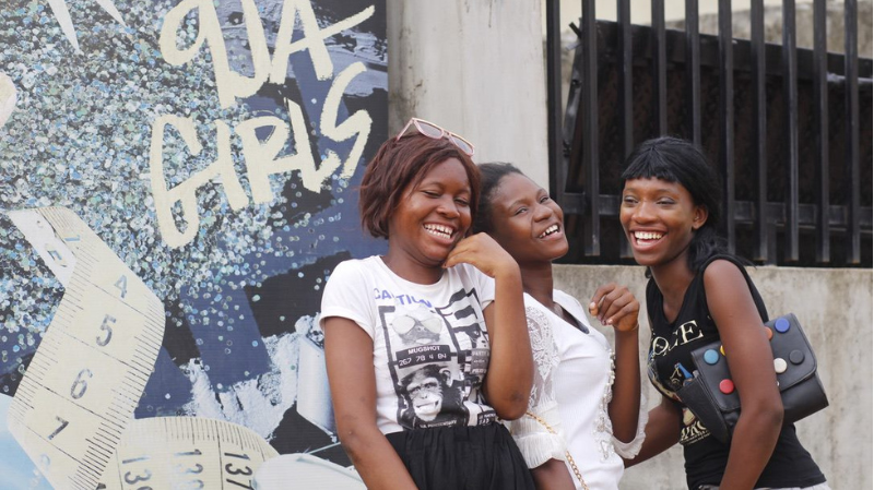In Nigeria, three girls laugh together in front of a billboard that says 'Girls'.