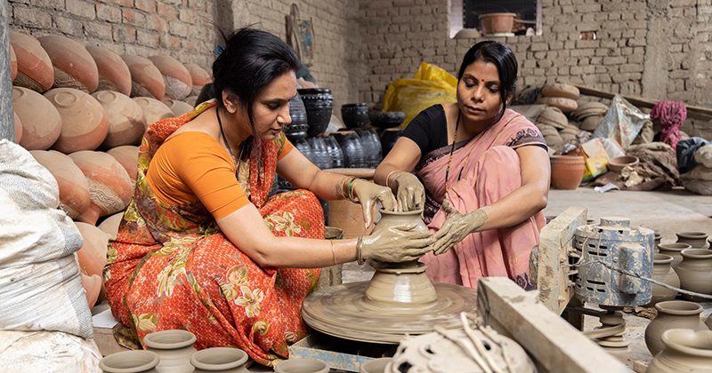 Two Indian women sit on the floor while sculpting pottery. There are already made pots surrounding them on the floor.