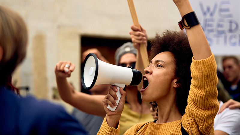 A Black women protesting, her arm is raised in the air and she is speaking into a megaphone. She is wearing a yellow jumper. A person behind her is holding a wooden signpost.