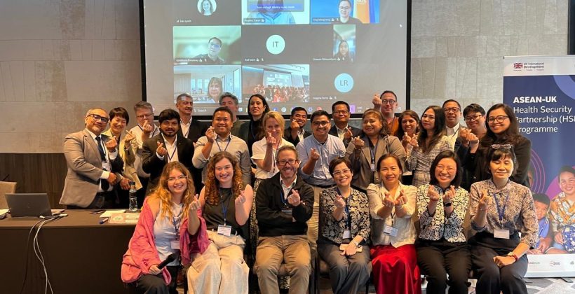 A diverse group of 26 participants of the learning forum pose for the camera in front of a large projector screen showing 9 online participants. They are smiling and clicking their fingers.